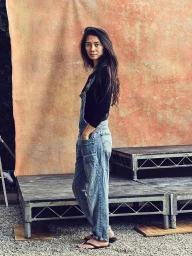 Woman wearing denim overalls and a black long-sleeve top standing on a platform against an orange mottled backdrop.