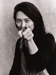Portrait of filmmaker Chloe Zhao pointing at the camera, wearing overalls over a black top against a textured wall.