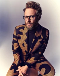 Portrait of actor Seth Rogen wearing a patterned suit and glasses, seated against a gradient backdrop.