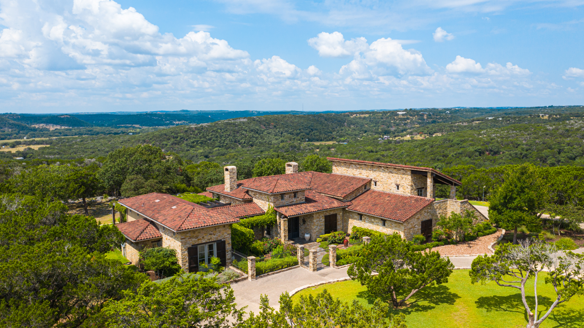 Wide shot of a large, light-stone house with a red tile roof, surrounded by trees, with rolling green hills in the background under blue sky.