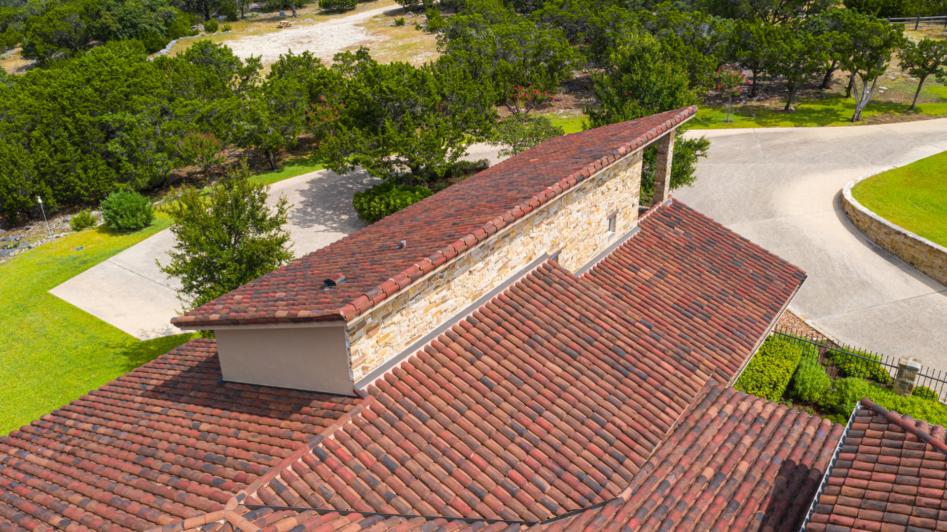 High-angle view of the roof of a house, showing terracotta-colored tiles and stone accents, with trees and greenery in the background.