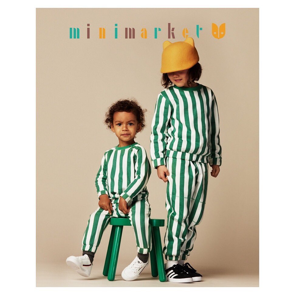Two young children in matching green and white striped outfits, one sitting on a green stool, in a studio shot with a neutral background.