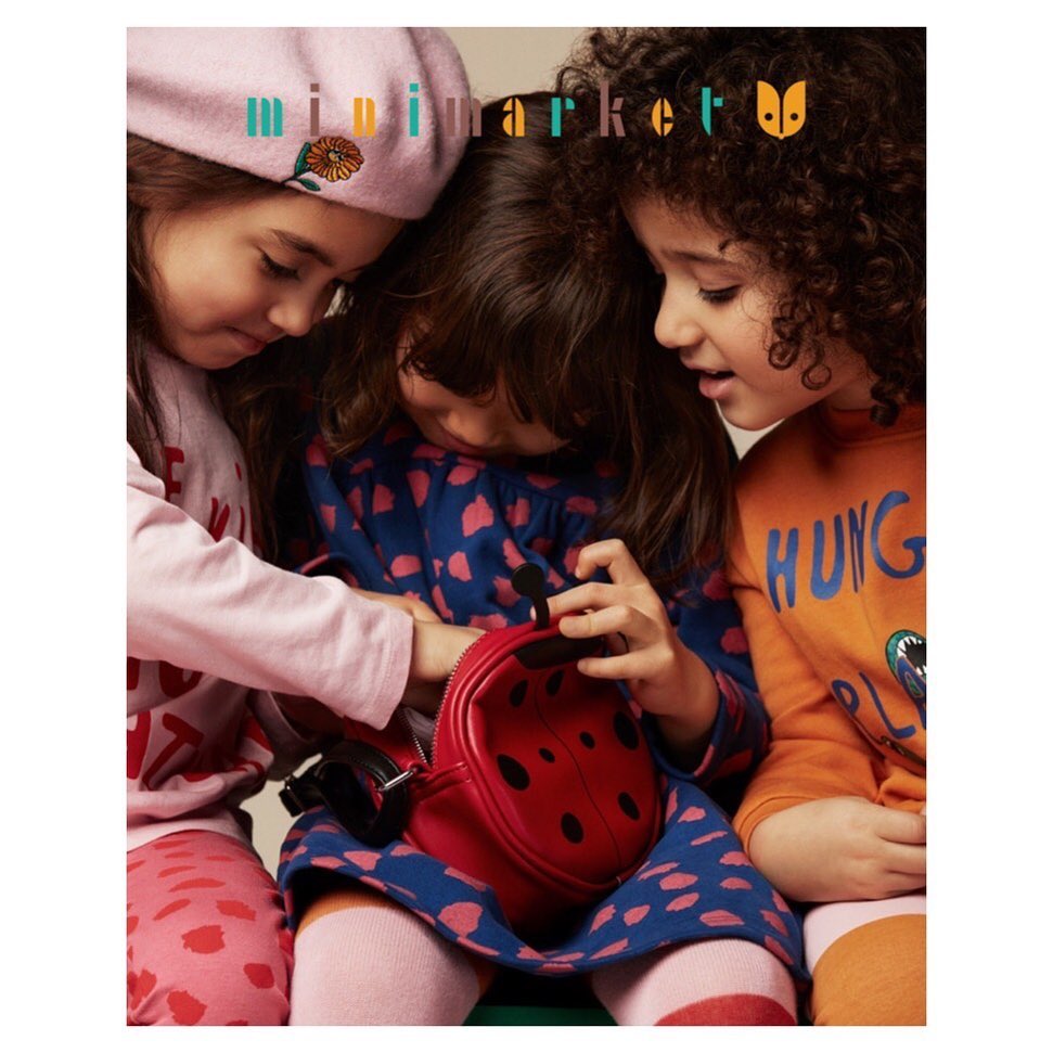 Three young girls in colorful patterned outfits huddle together; the girl in the middle is holding a red ladybug-shaped purse.