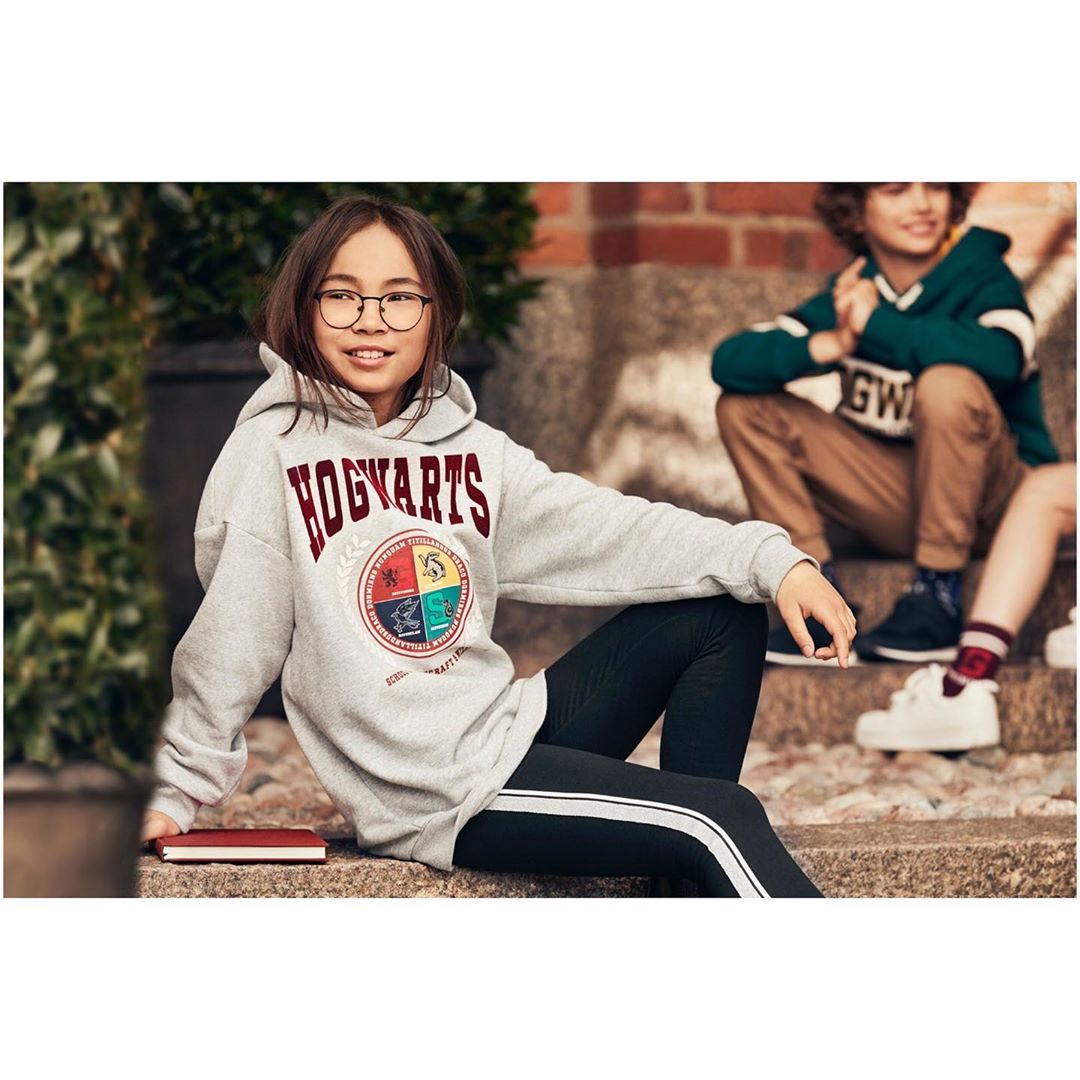 A young girl wearing a gray Hogwarts hoodie sitting on steps outside, with a boy in the background, and a red book beside her.