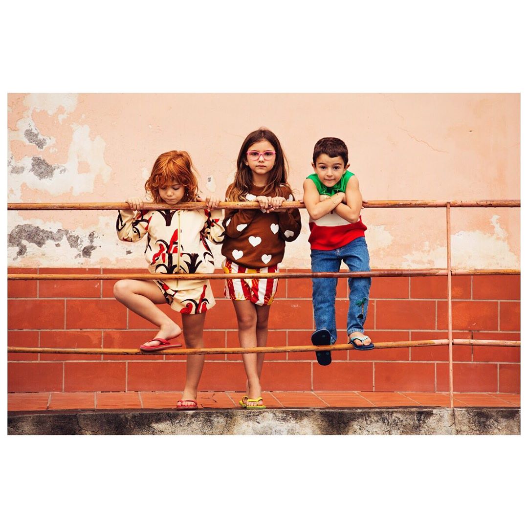 Three children stand against a peach-colored wall and a red tile base, posing on a rusty metal rail in colorful clothes with unique patterns.