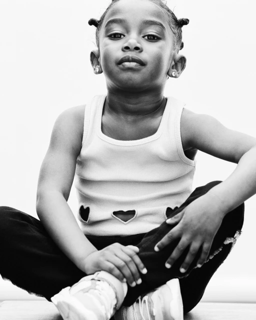 Portrait of a young Black girl with a serious expression, wearing a light tank top and dark pants, sitting cross-legged against a white background.