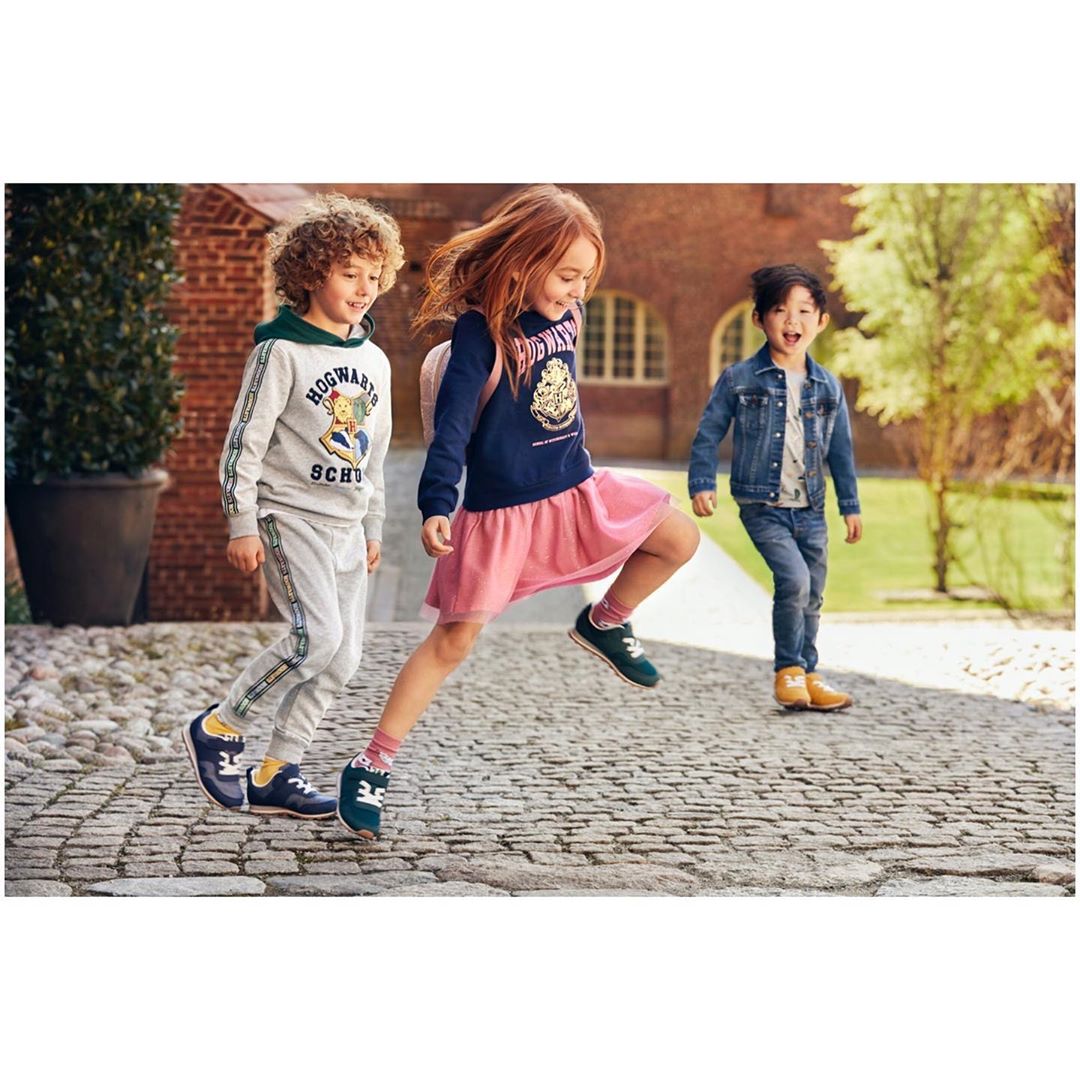 Three kids in Harry Potter sweatshirts jumping on a brick path outdoors, with trees and a building visible in the background.