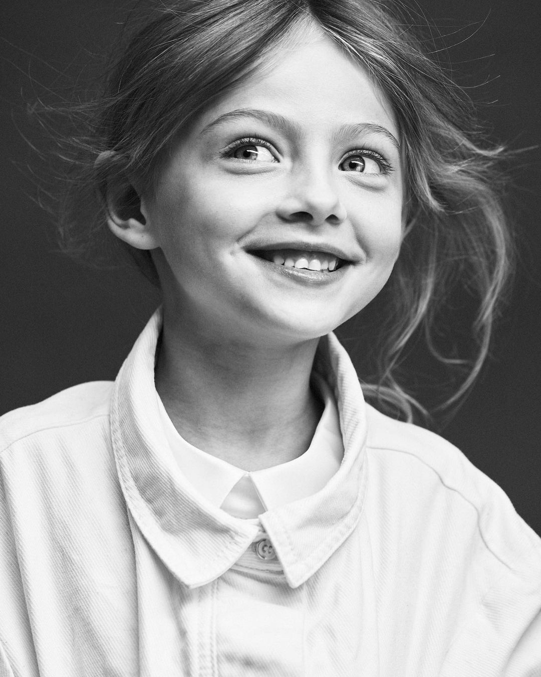 Portrait of a young girl with light hair smiling in a black and white studio shot, wearing a jacket and collared shirt.