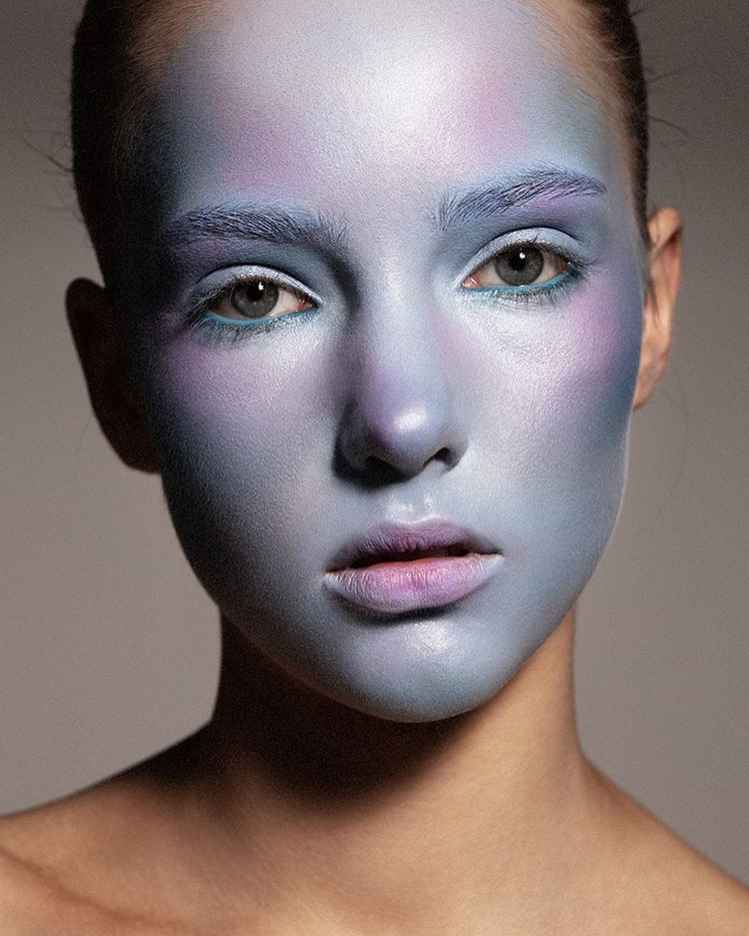 Close-up of a woman with light skin and blue and purple makeup covering her face and eyebrows in a beauty and fashion style portrait.