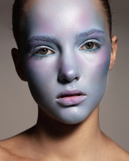 Close-up of a woman with light skin and blue and purple makeup covering her face and eyebrows in a beauty and fashion style portrait.
