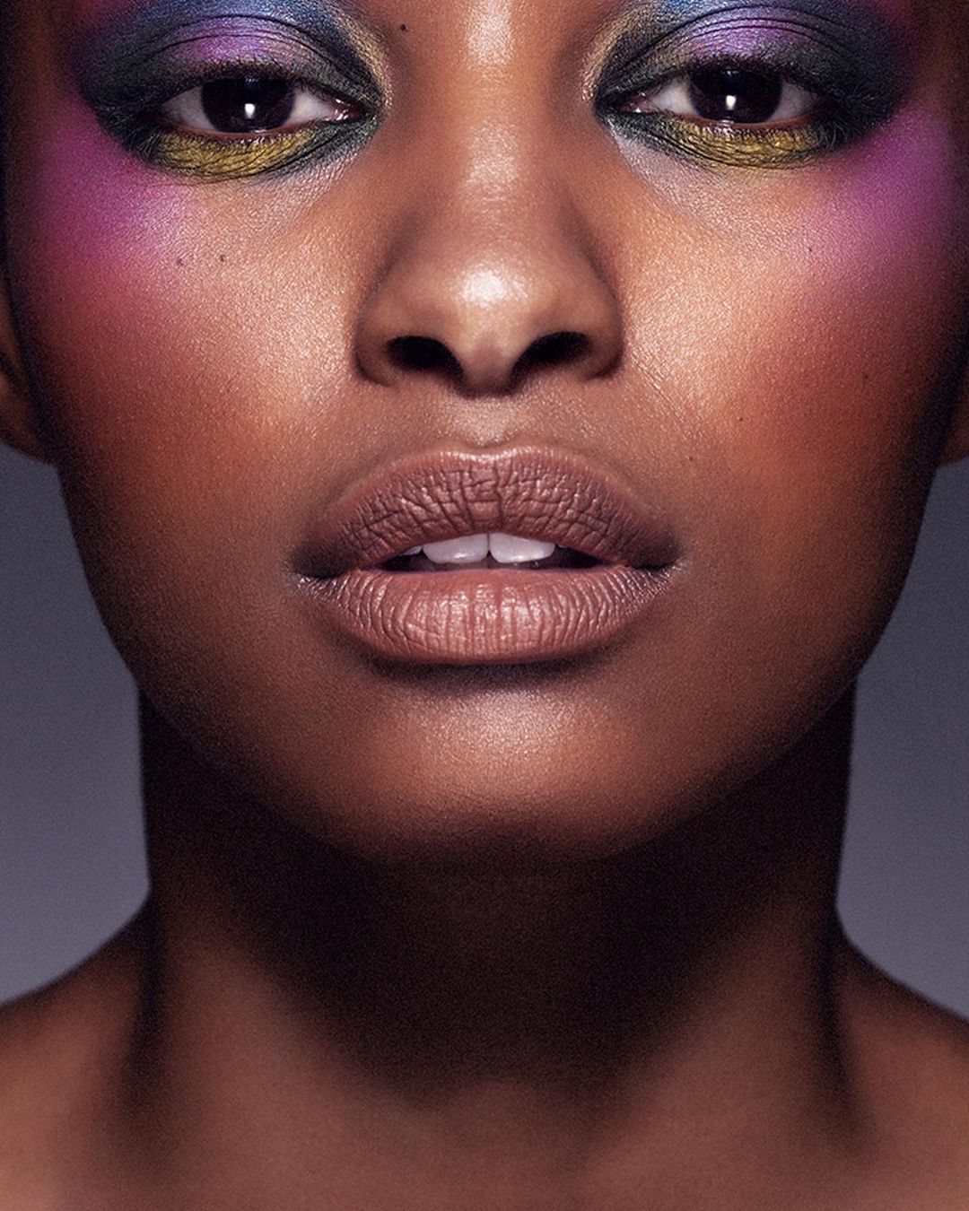 Close-up beauty shot of a Black female adult model with purple, blue, and gold eye shadow, light pink lipstick, and shiny skin.