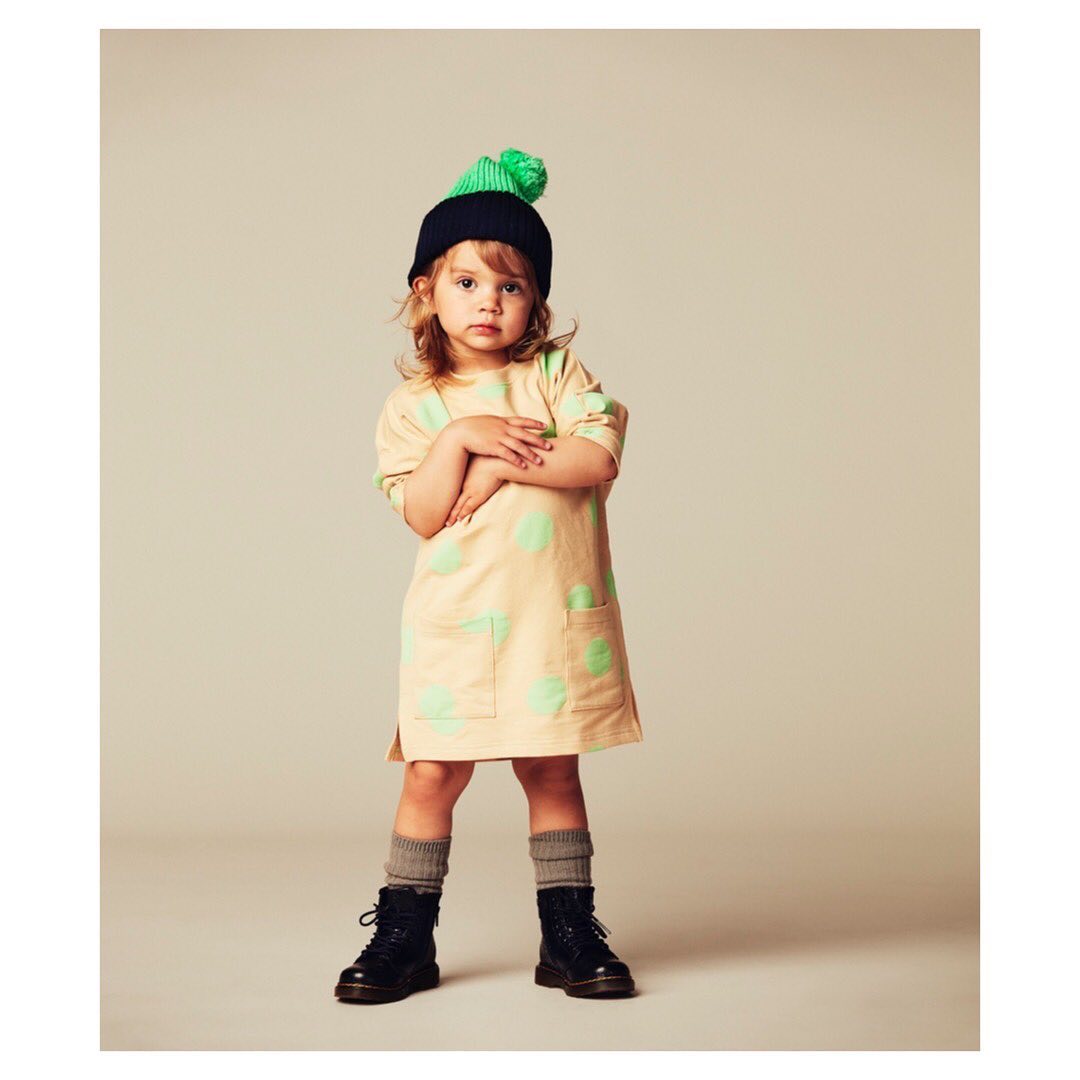 Young child in a polka dot dress, black boots, and a green and black beanie, standing in a neutral studio setting, looking at the camera.