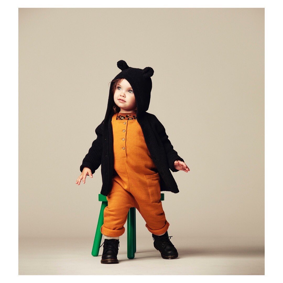 Adorable toddler in an orange jumpsuit and black bear-eared hoodie, standing on a green stool in a studio setting.