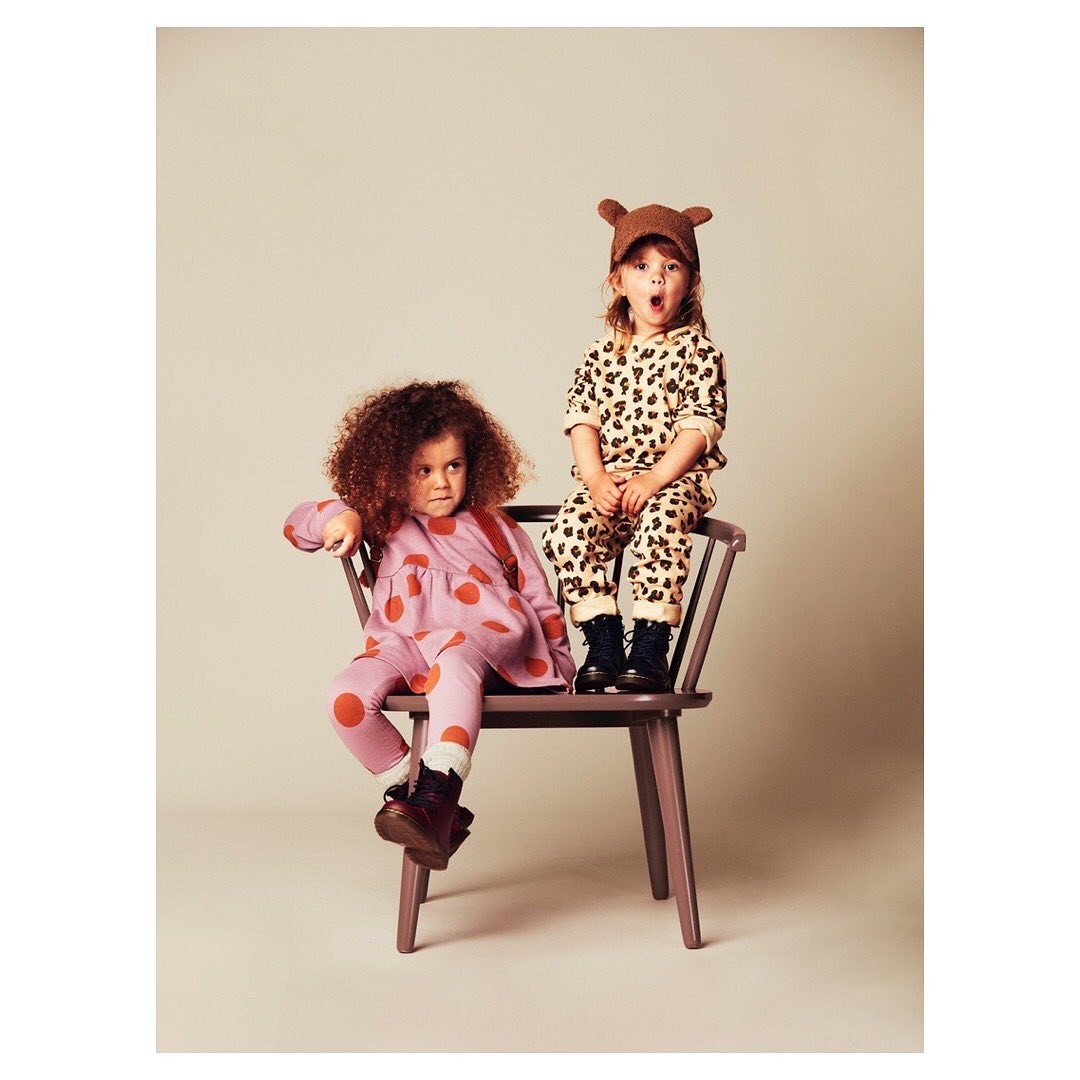 Two young children sit on a wooden chair, one in a leopard print outfit and the other in a lilac polka dot dress and leggings.