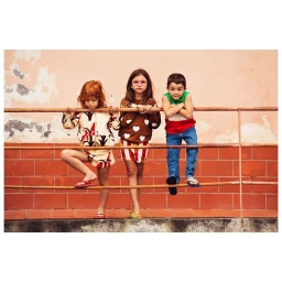 Three children leaning against a metal railing in front of a textured pink and tiled wall by Joel Rhodin.