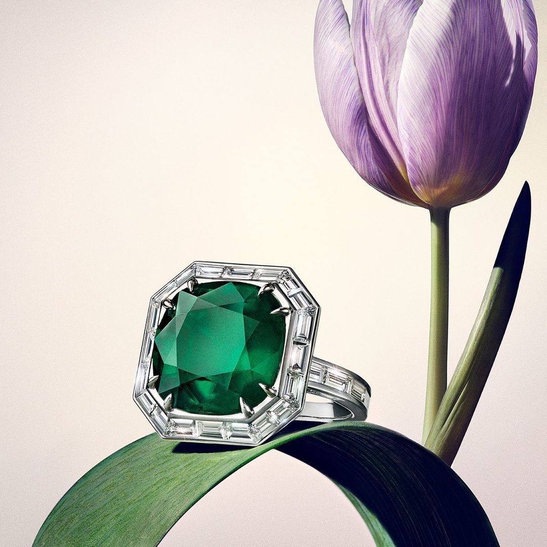 Elegant emerald-cut emerald ring with diamond accents, resting on a green leaf with a blurred tulip in the background. Jewelry still life.