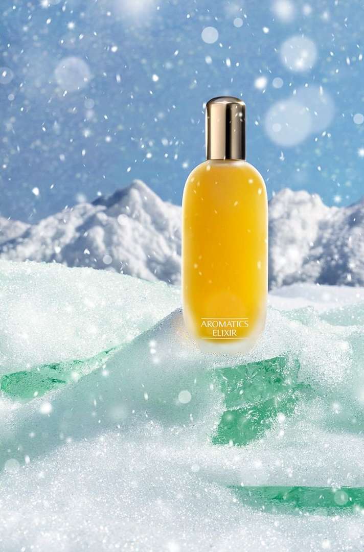 Aromatics Elixir bottle sits on snowy ice with a mountain backdrop and bokeh snow effects in a beauty and personal care advertisement.