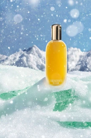 Aromatics Elixir bottle sits on snowy ice with a mountain backdrop and bokeh snow effects in a beauty and personal care advertisement.