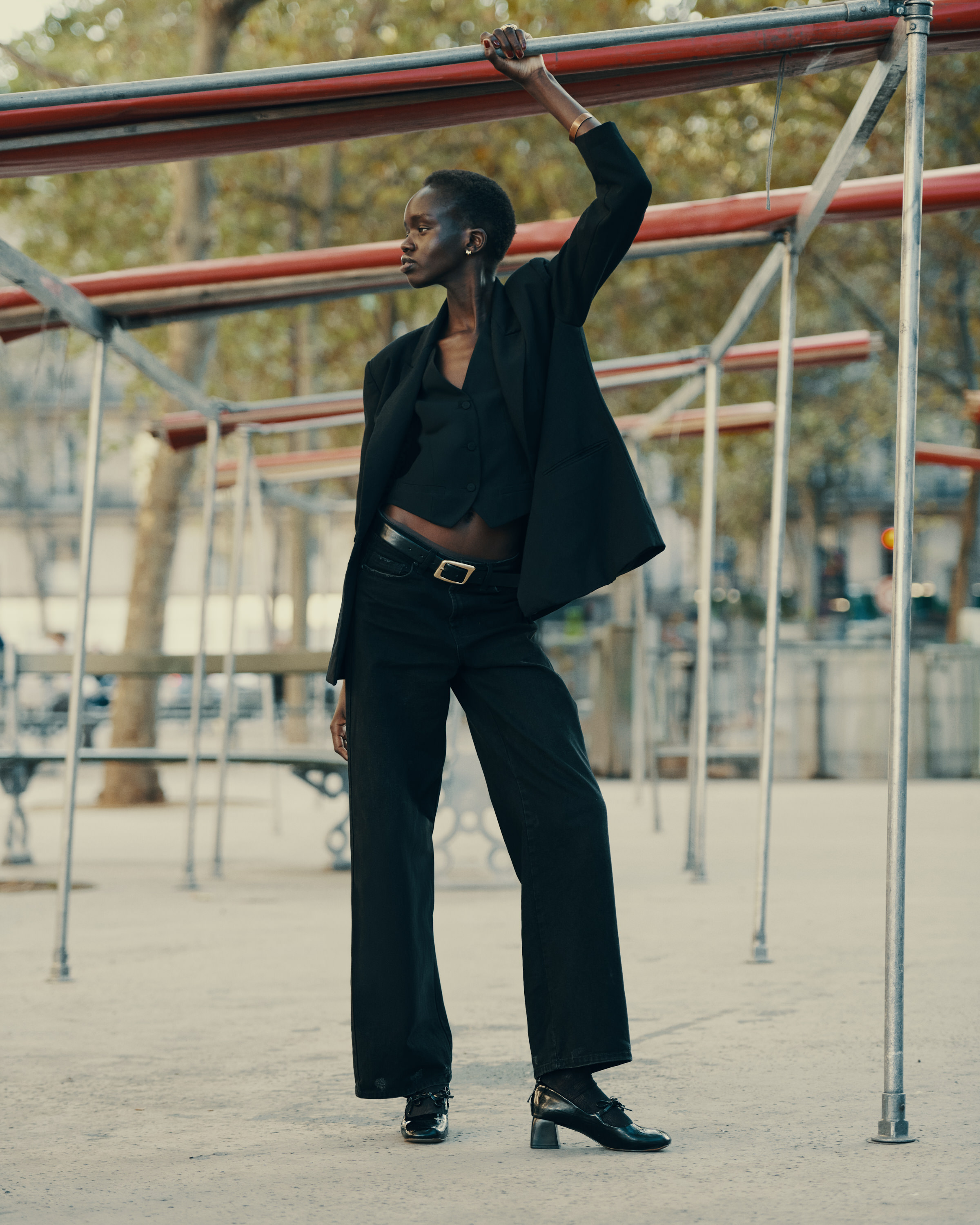 A slender African-American woman poses outdoors in black blazer, vest, and wide-legged pants, holding onto a metal structure on a sunny day.