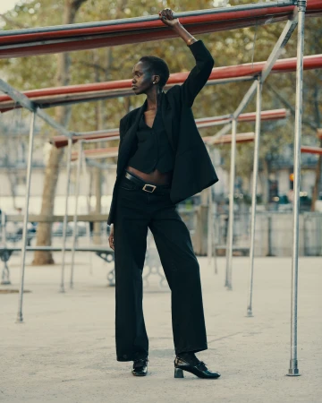 A slender African-American woman poses outdoors in black blazer, vest, and wide-legged pants, holding onto a metal structure on a sunny day.