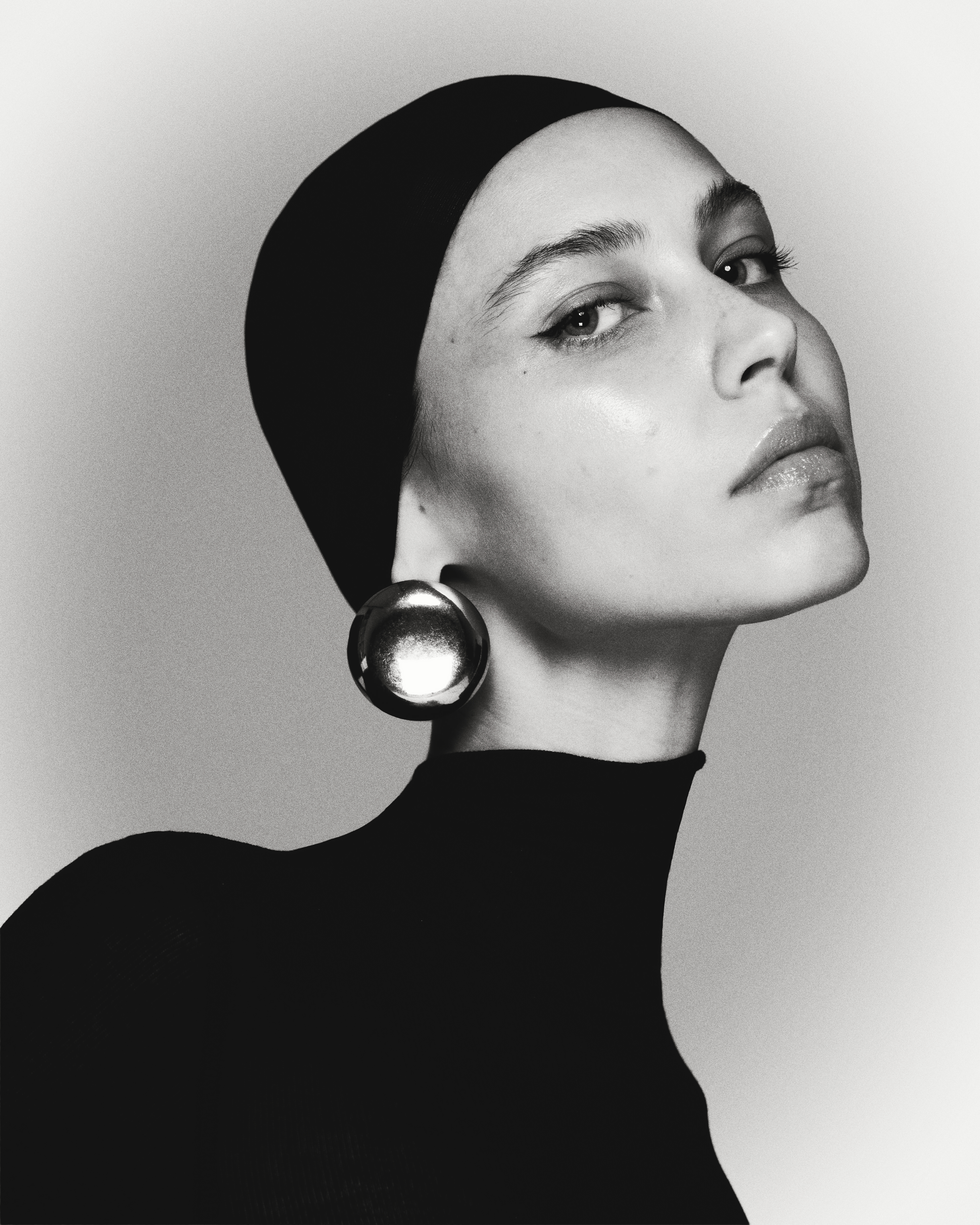 Elegant close-up portrait of a young woman wearing a black headscarf, black turtleneck top, and large, round, reflective earrings. Studio lighting.