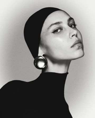 Elegant close-up portrait of a young woman wearing a black headscarf, black turtleneck top, and large, round, reflective earrings. Studio lighting.