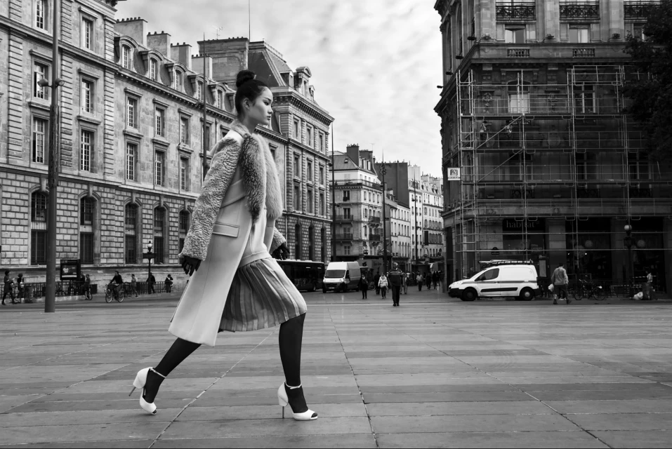 Fashion model in a fur-collared coat walks across a stone plaza in a black and white shot, framed by ornate buildings and city streets.