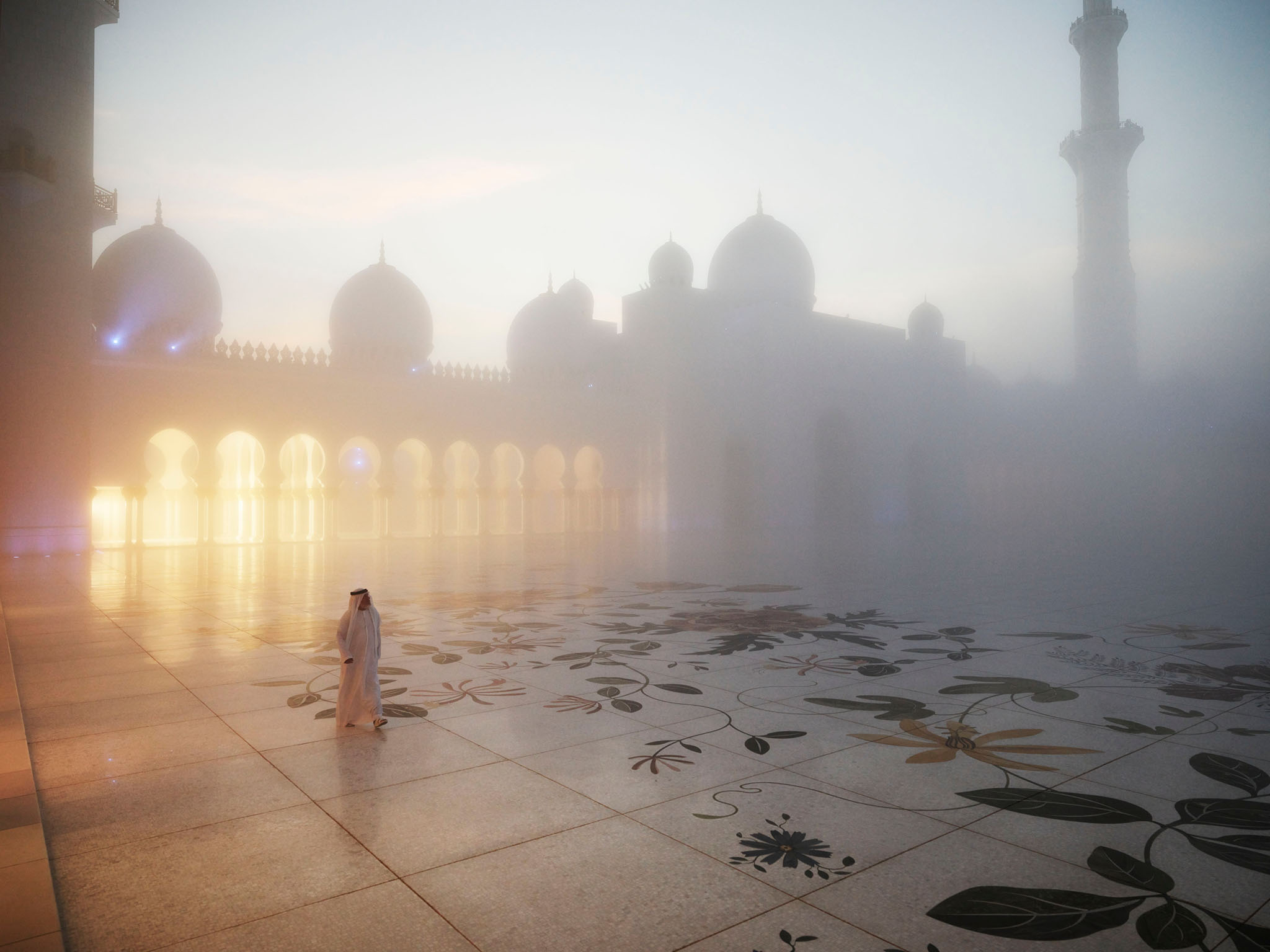 A man in traditional Gulf attire walks across a marble plaza in front of a mosque in the United Arab Emirates under foggy conditions.