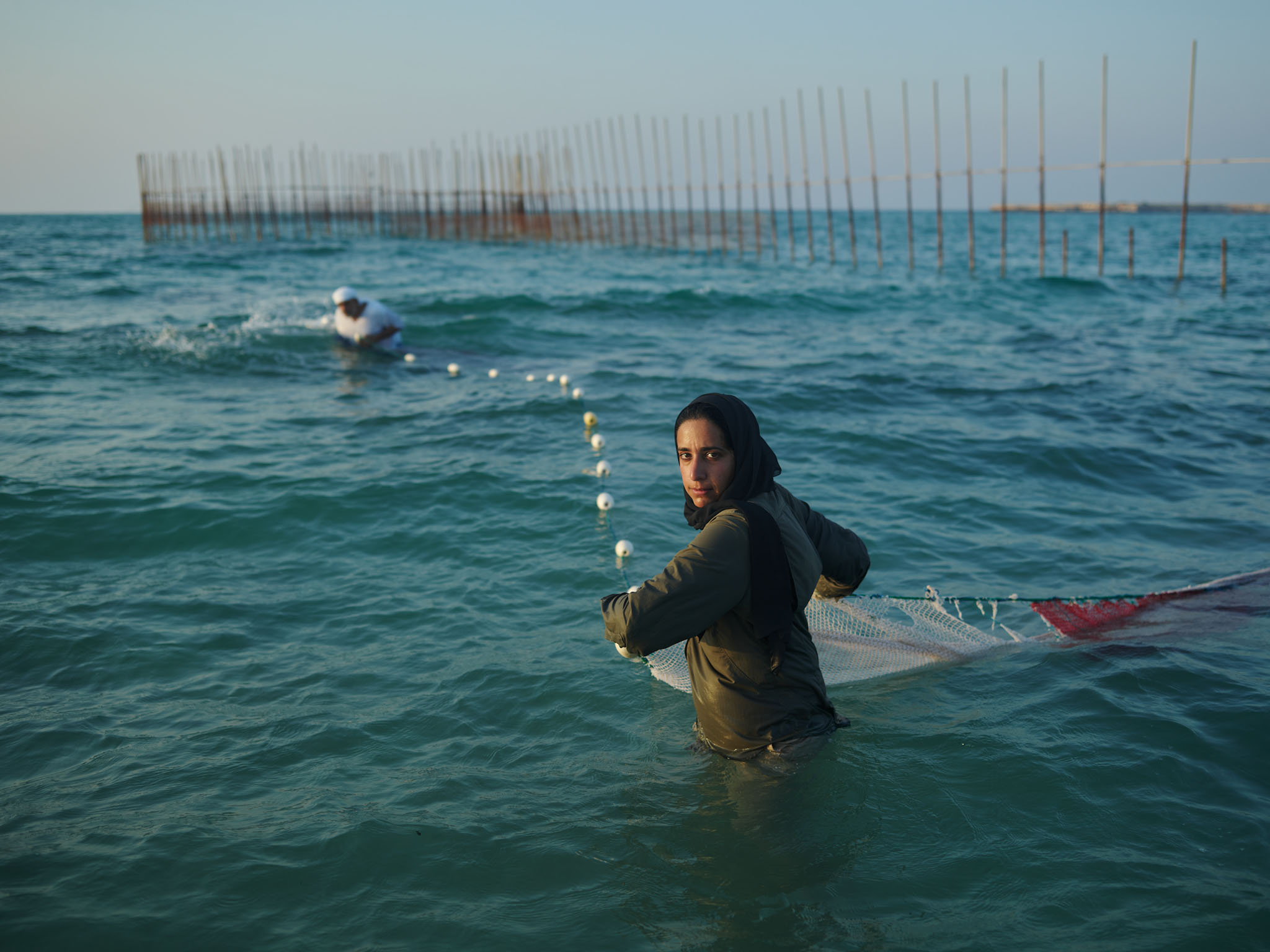 Woman in headscarf stands in ocean holding net with other person in background at fishing weir in teal-colored water under blue sky.