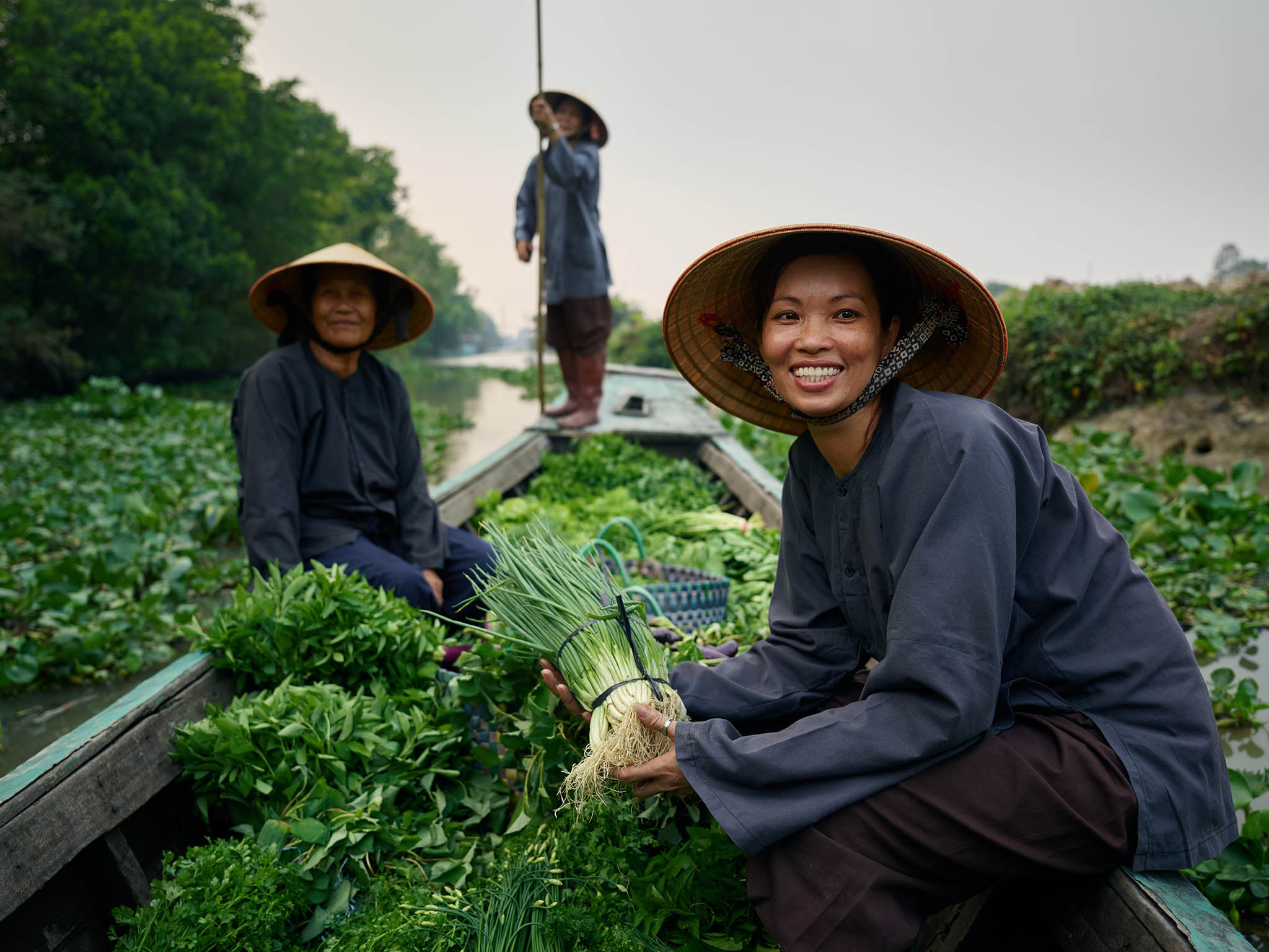Three women in conical hats on a boat laden with fresh produce on a waterway, smiling and engaging the viewer in a rural setting.