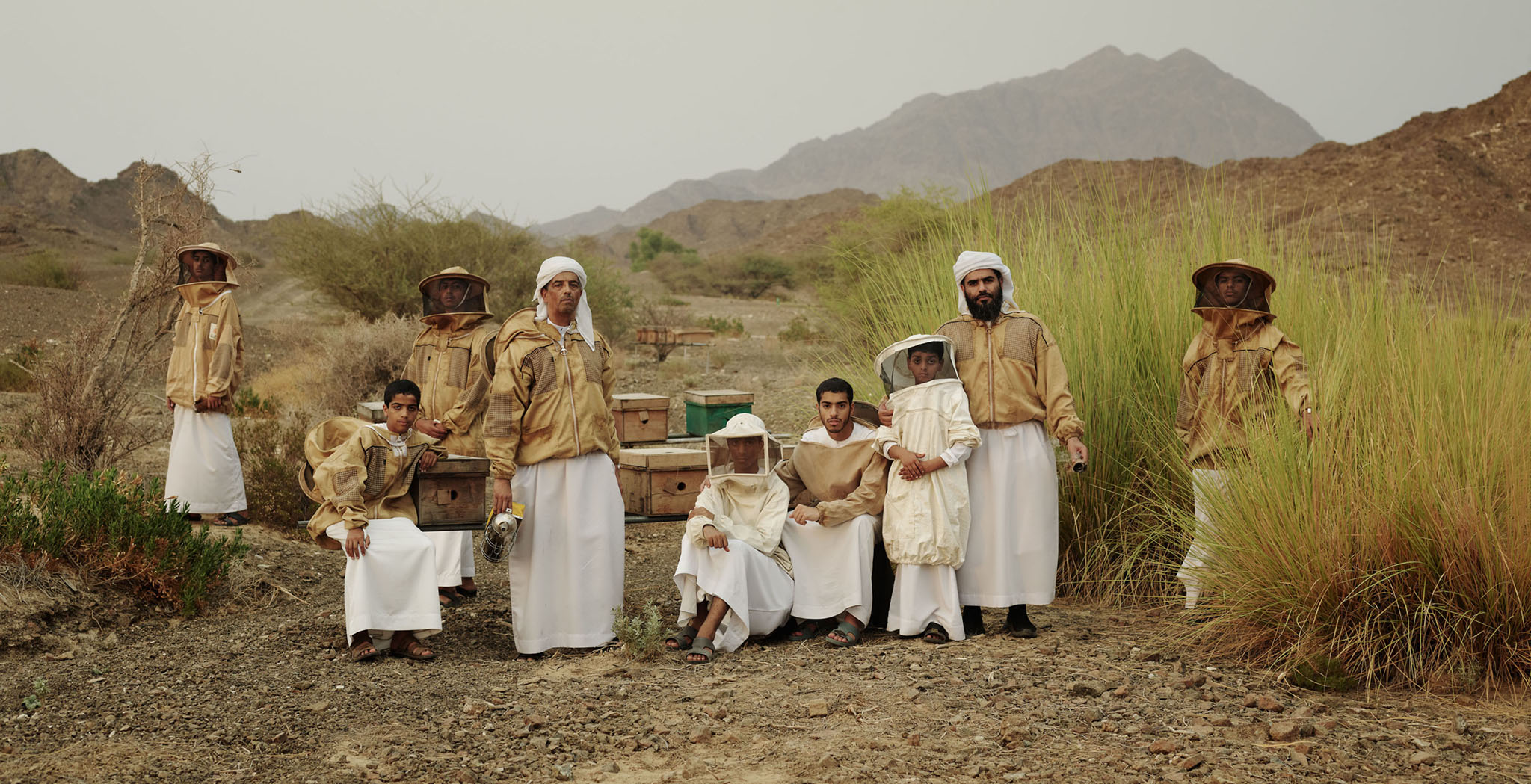 Group of beekeepers in protective suits and traditional clothing stand near beehives in a rural, rocky, mountainous landscape.