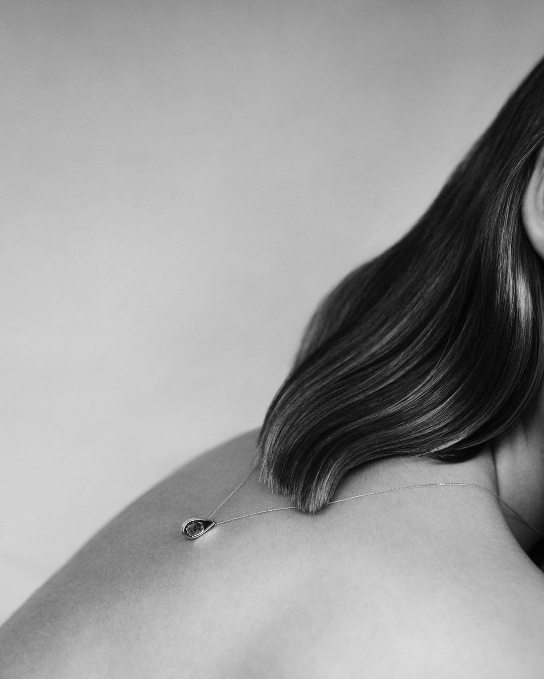 Close-up black and white photo of a woman's bare shoulder and the back of her neck, wearing a teardrop pendant necklace.
