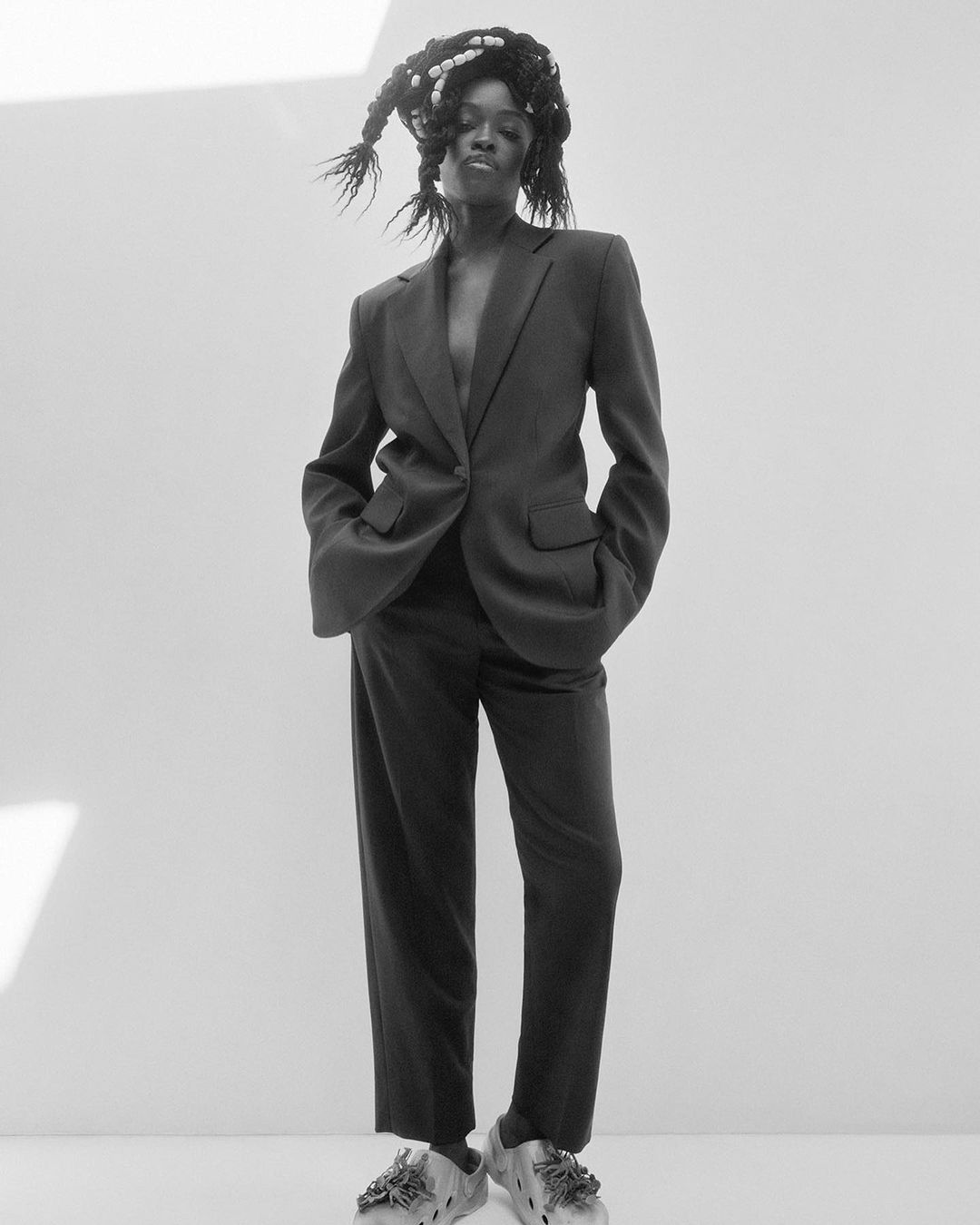 Monochrome full shot of androgynous adult in dark suit with braided hair, standing in photo studio with hands in pockets and white background.
