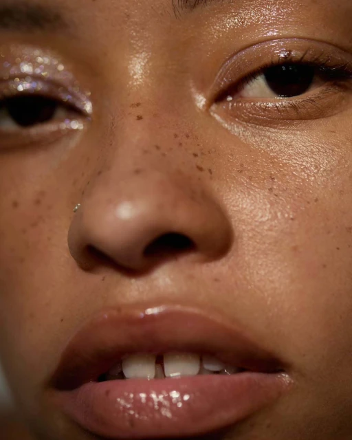 Close-up portrait of a young adult female with highlighted cheekbones, glossy lips, freckles, and glittery eye shadow looking at the camera.