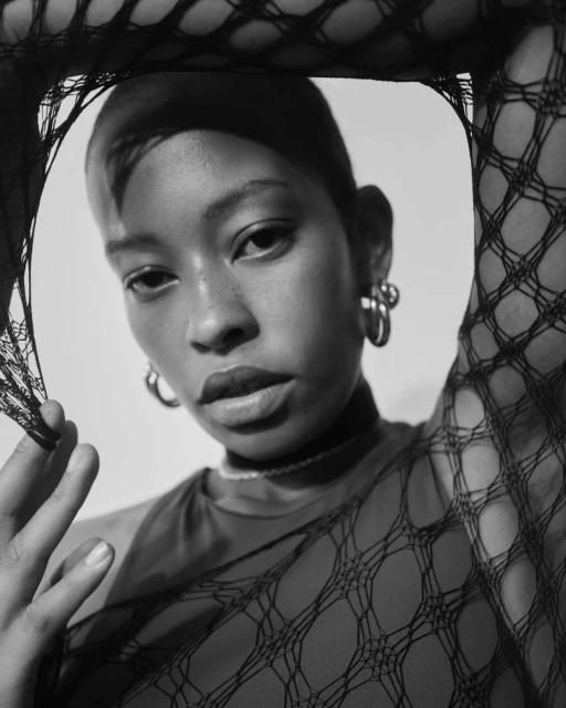 Monochrome portrait of a young woman with short hair wearing jewelry and a netted top against a light background.