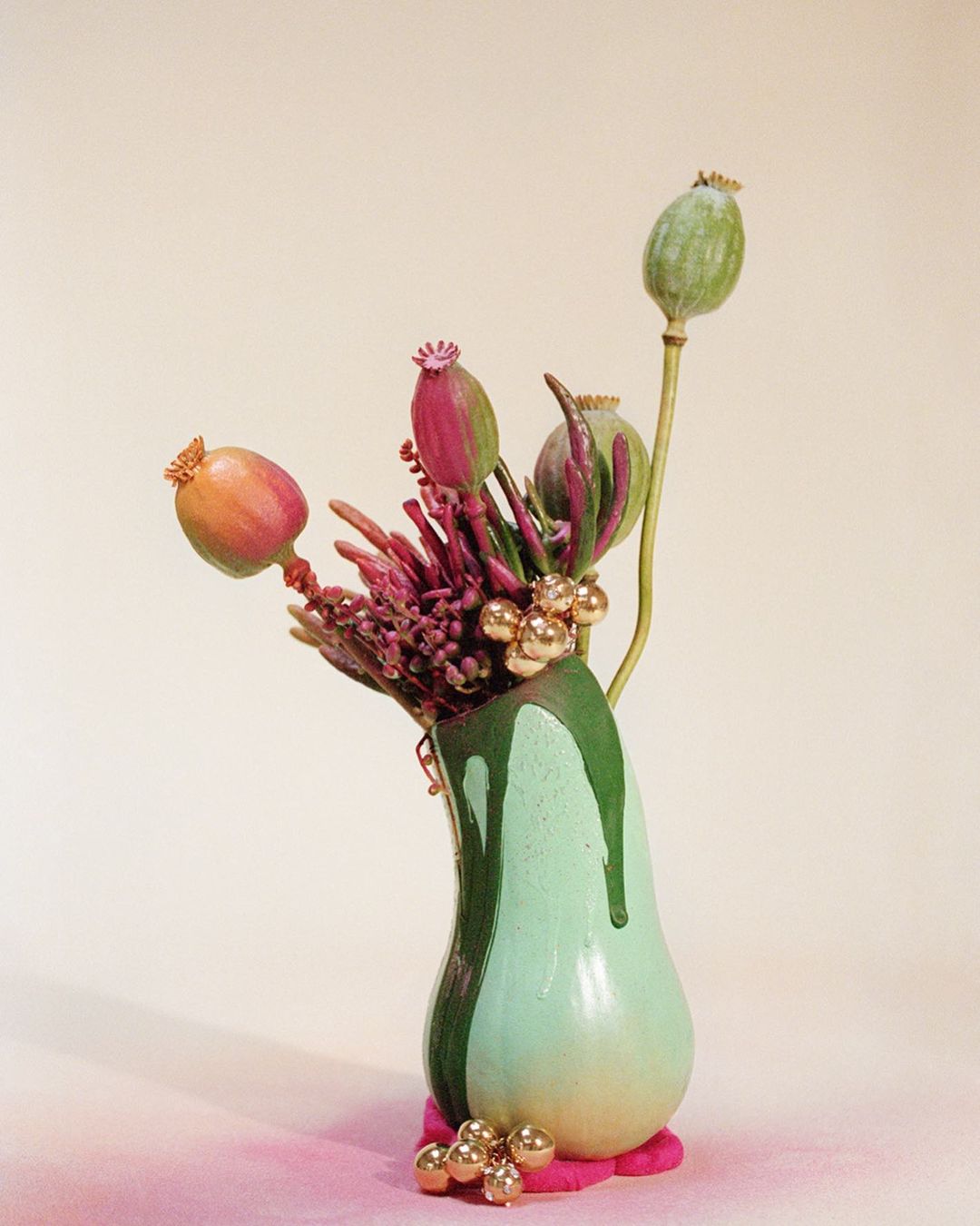 Still life arrangement featuring poppy seed pods and purple flowers in a green vase with gold accents on a light beige background.