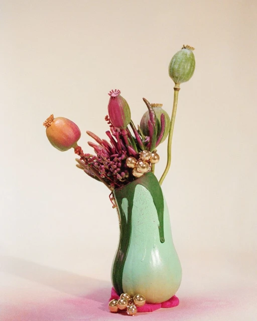 Still life arrangement featuring poppy seed pods and purple flowers in a green vase with gold accents on a light beige background.