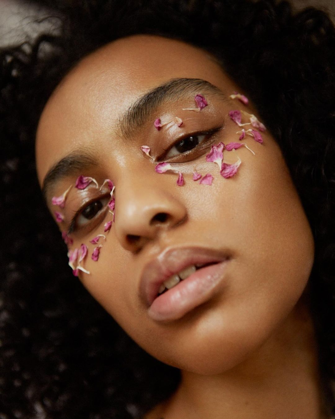 Portrait of a Black woman with flower petals around her eyes in a close-up shot. Soft focus showcases her skin and curly hair.