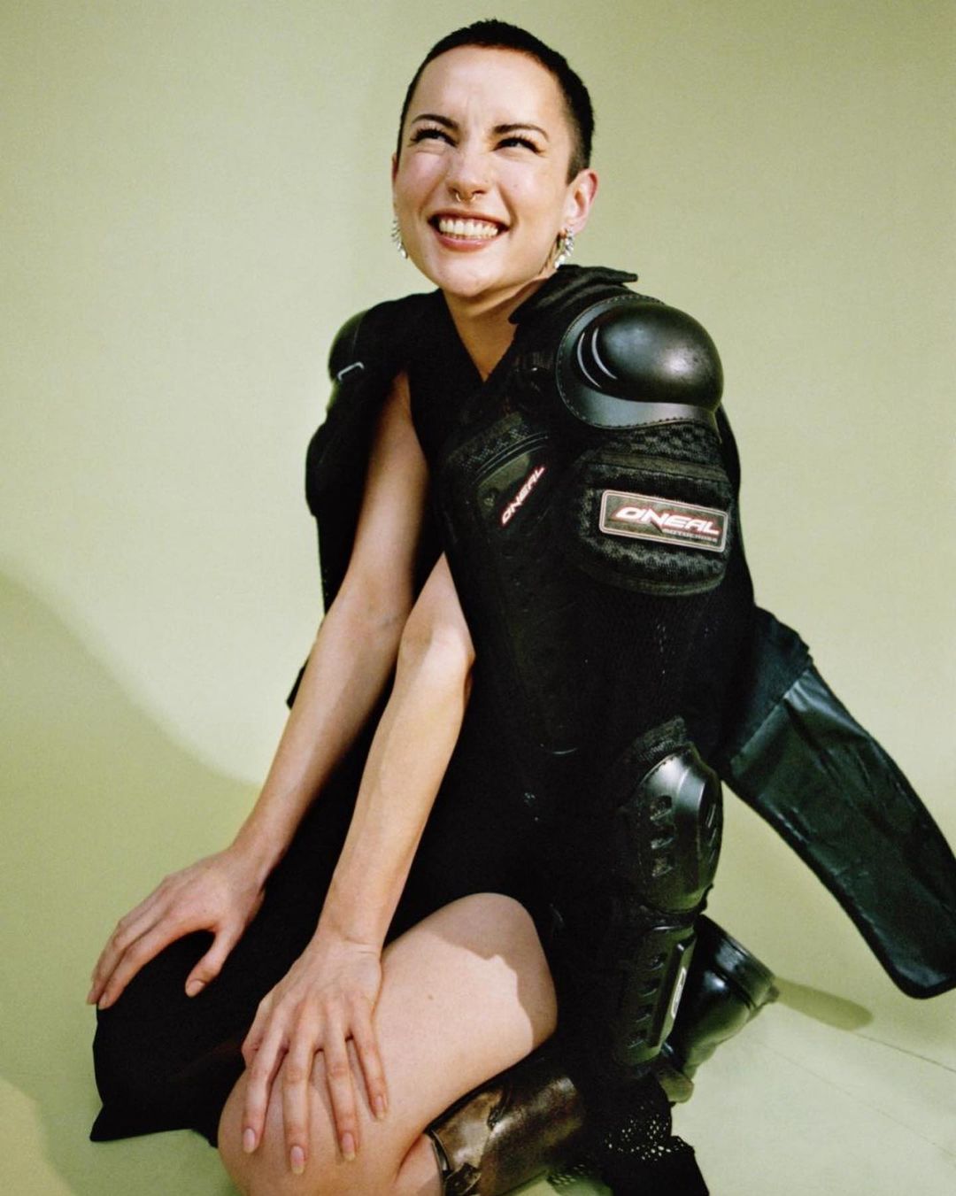 A smiling woman with a buzzcut poses in a black dress and black knee and shoulder pads against a light green backdrop in a studio.