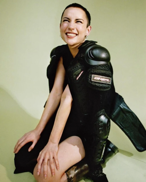 A smiling woman with a buzzcut poses in a black dress and black knee and shoulder pads against a light green backdrop in a studio.