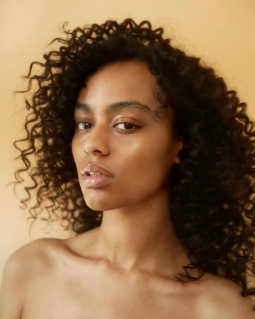 Close-up portrait of a woman with dark curly hair and glossy lips against a neutral warm background by Johanna Nyholm