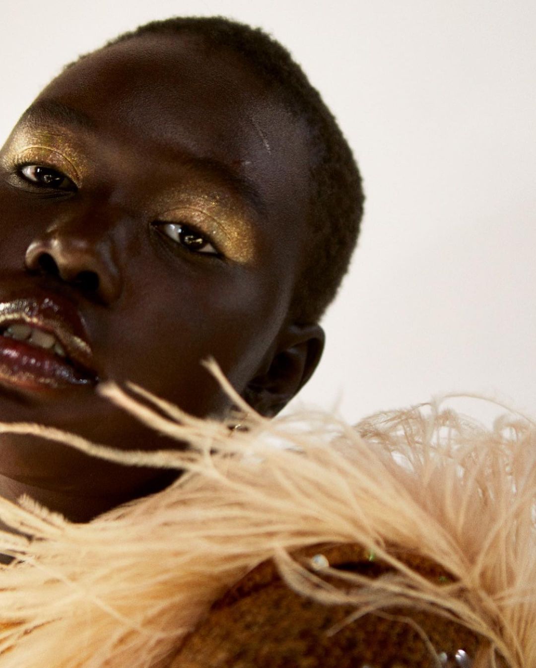 Close-up portrait of a woman with dark skin and golden makeup wearing a feathered garment against a neutral background.