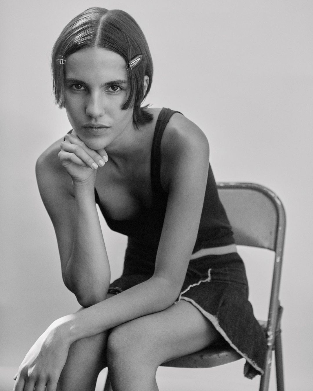 Black and white portrait of a young adult woman with hand to chin, wearing a dark tank top and skirt, seated on a metal chair.