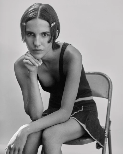 Black and white portrait of a young adult woman with hand to chin, wearing a dark tank top and skirt, seated on a metal chair.