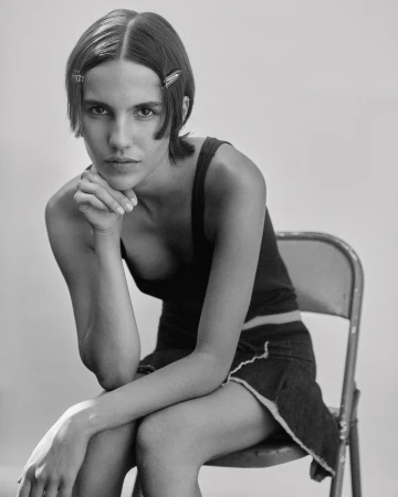 Black and white portrait of a young adult woman with hand to chin, wearing a dark tank top and skirt, seated on a metal chair.