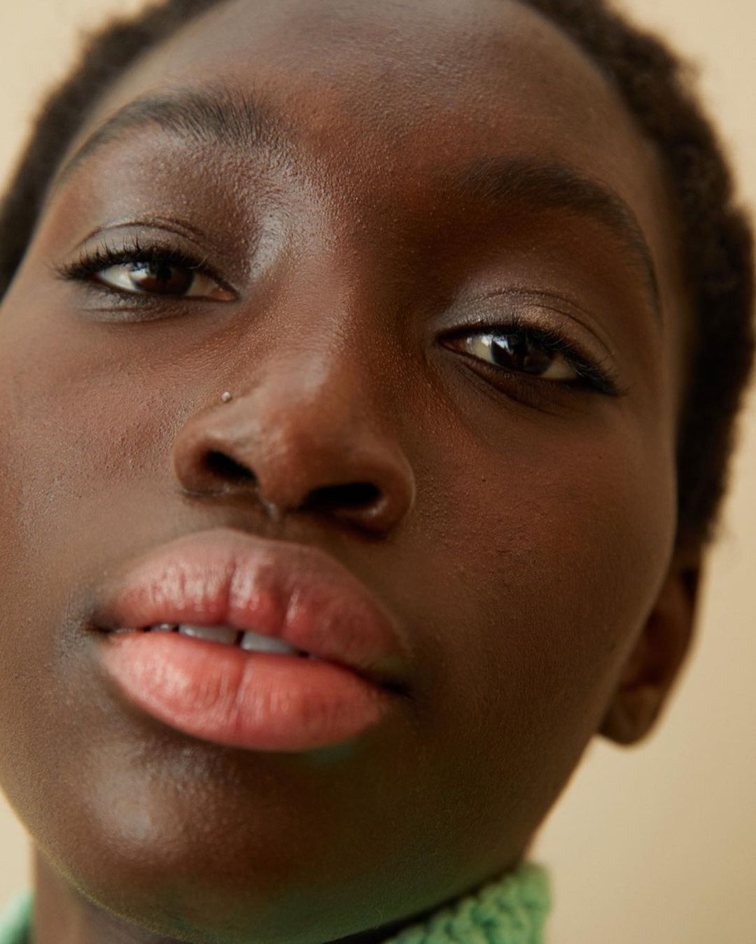 Close-up portrait of a dark-skinned woman with soft, neutral makeup and a light green knit collar, set against a neutral beige backdrop.