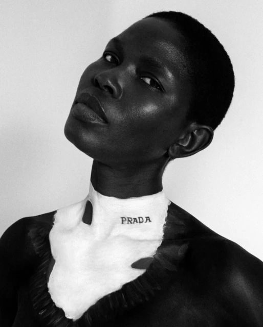 Close-up portrait of a dark-skinned woman with short hair, wearing white body paint with the PRADA logo on her neck in black and white.