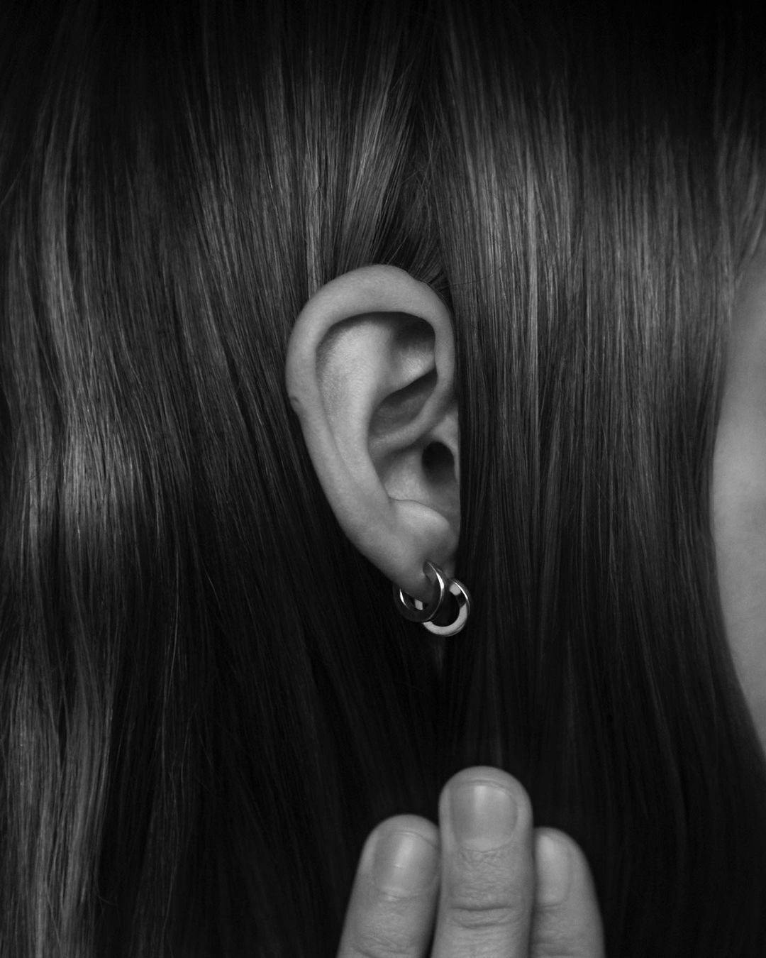 Close-up shot in monochrome of a young adult female's ear with silver hoop earrings and hair. The fingers are visible.
