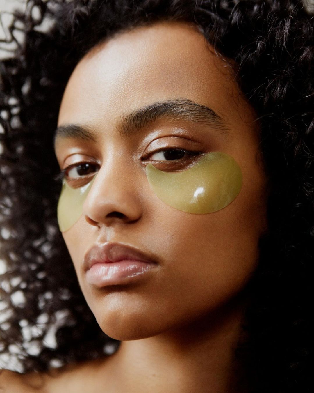 Close-up of adult female with olive-green under-eye patches on light brown skin, curly dark hair, neutral expression, beauty concept.