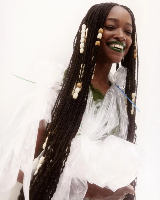 A fashionable, young, Black woman smiles brightly with green lipstick and beaded braids, wearing a plastic wrap dress against a bright white background.